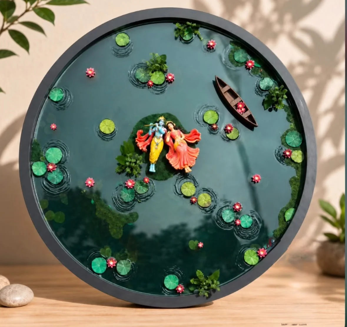 Circular resin art piece depicting Radha and Krishna standing on an island in a pond with lotus leaves, pink flowers, and a s