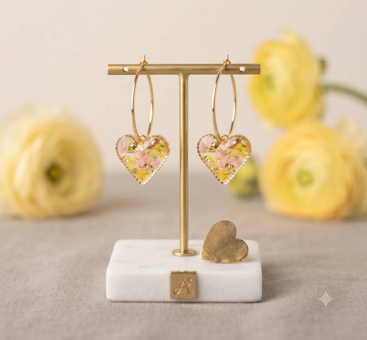 Pair of clear resin heart hoop earrings with tiny pink and yellow dried flowers, displayed on a gold stand.