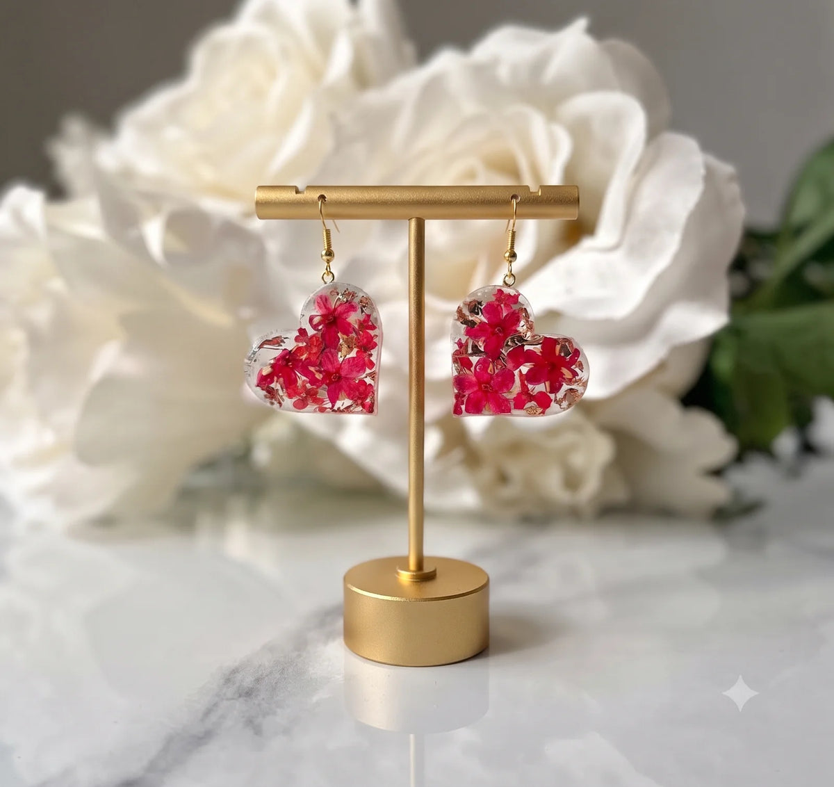Pair of clear heart-shaped resin earrings with vibrant red dried flowers and gold flakes, displayed on a gold stand.