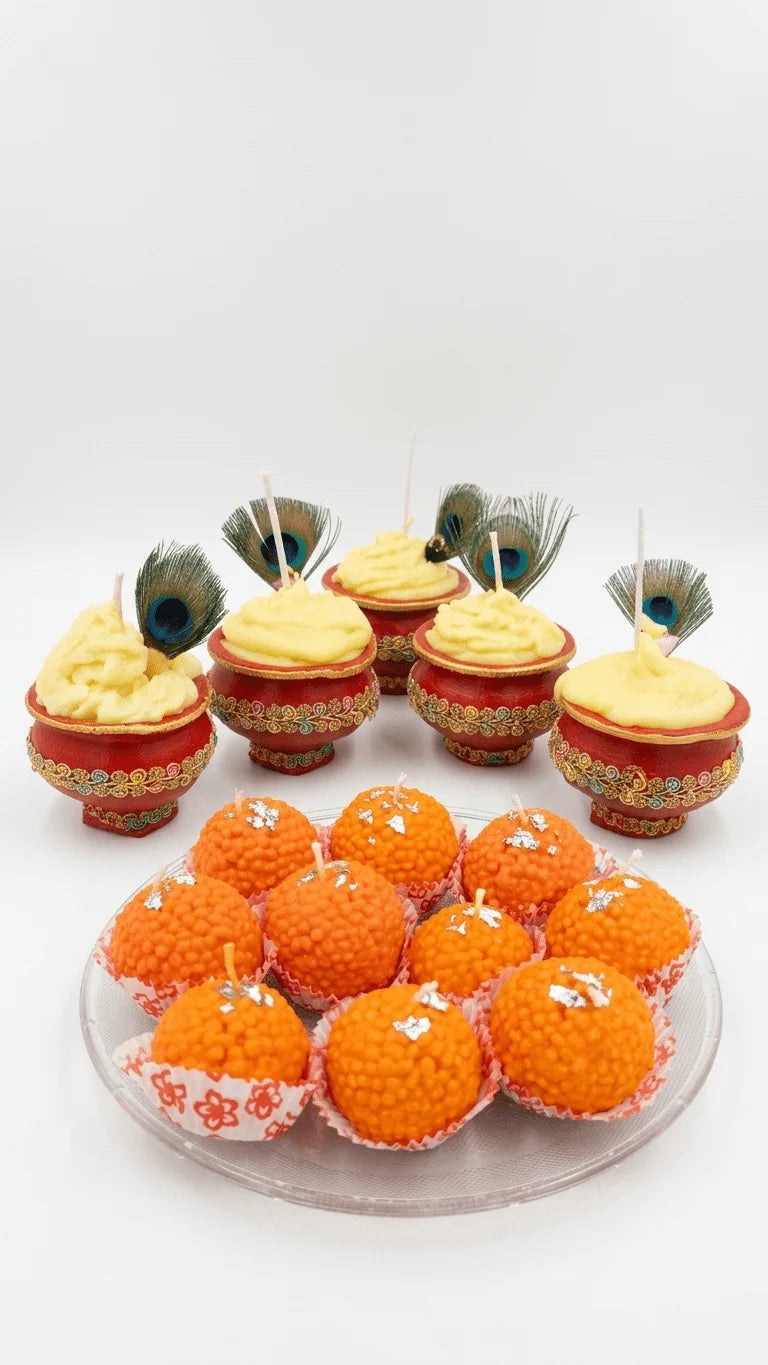 Red pots with yellow candles and peacock feathers, alongside orange ladoo-shaped candles with silver leaf on a clear plate.