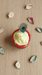 Cream swirled candle in a red decorated diya with a peacock feather, surrounded by dried leaves on a wooden surface.