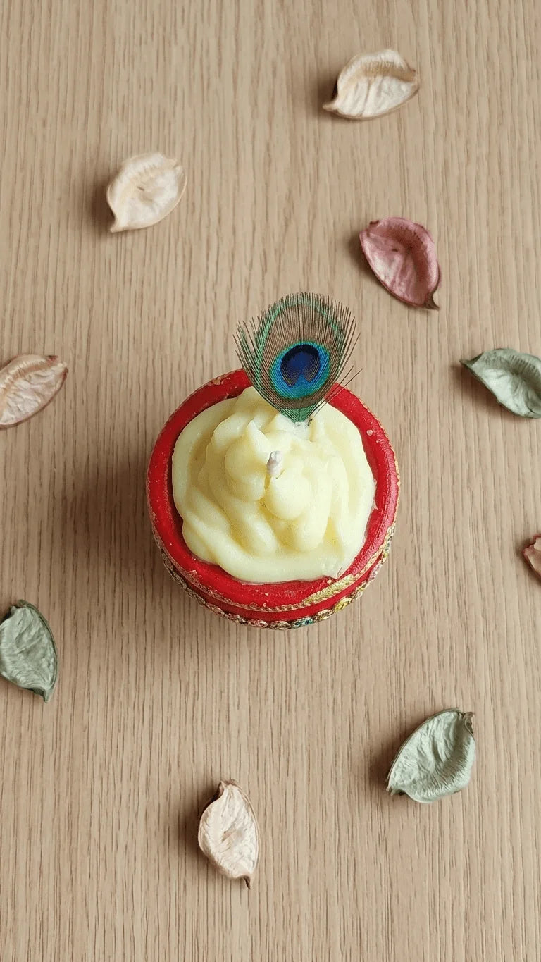 Cream swirled candle in a red decorated diya with a peacock feather, surrounded by dried leaves on a wooden surface.