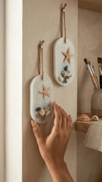 Two oval resin keepsakes with starfish, shells, and glitter hang on a wall, one being adjusted by a hand.