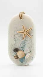 Oval white wax sachet adorned with a starfish, assorted seashells, sea glass, and teal glitter, with a jute string hanger.