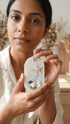 Woman holding a white oval resin keepsake decorated with a starfish, small shells, and blue glitter, against a soft backgroun