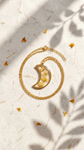 Gold chain necklace with a crescent moon resin pendant containing yellow dried flowers, on textured paper with leaf shadows.