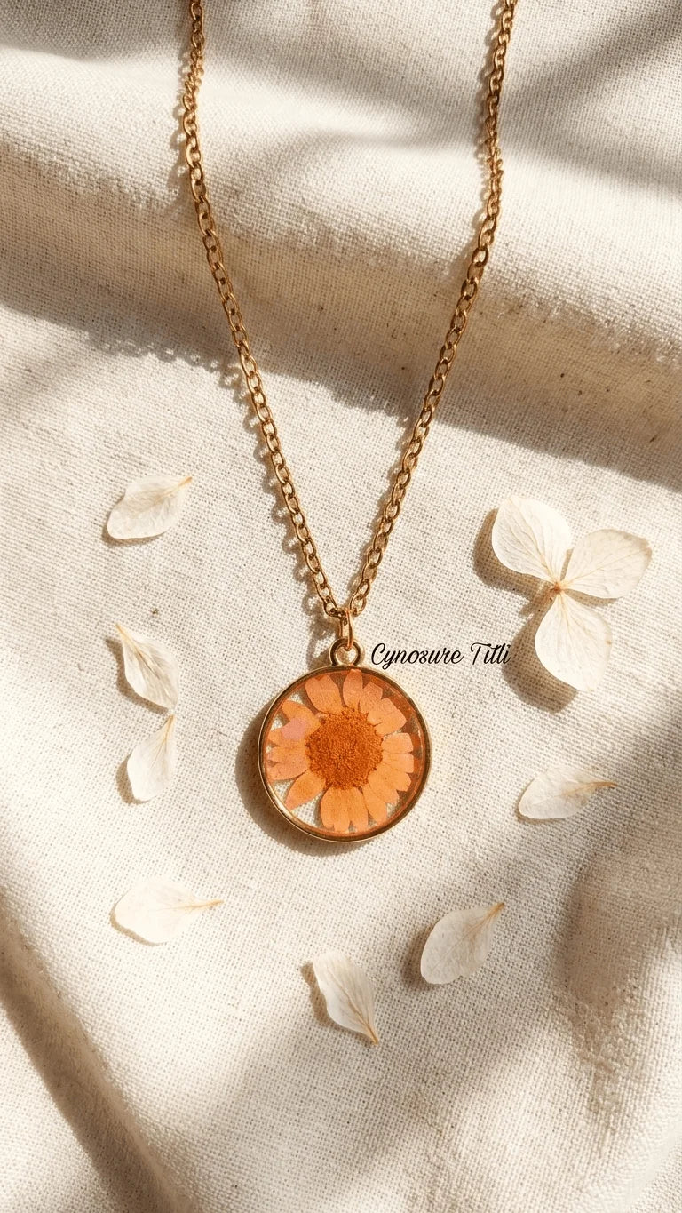 Gold chain necklace with a round resin pendant featuring an orange flower, laid on a light fabric with scattered petals.