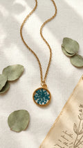 Gold chain necklace with a round resin pendant featuring blue dried flowers, on a white fabric with eucalyptus leaves and old
