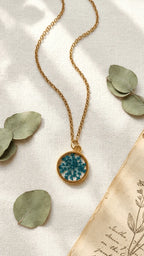 Gold chain necklace with a round resin pendant featuring blue dried flowers, on a white fabric with eucalyptus leaves and old