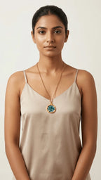 Woman wearing a beige camisole and a long gold chain with a round teal and white patterned resin pendant.