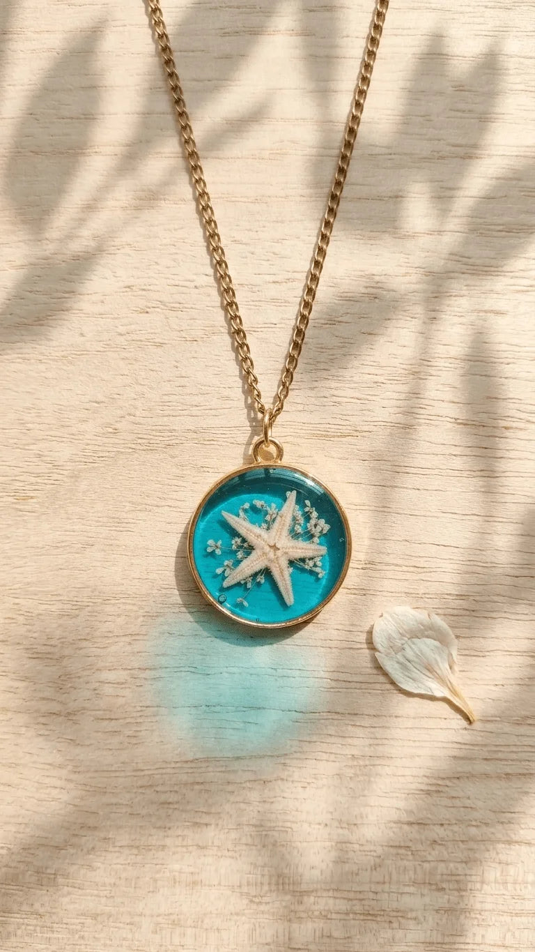 Gold chain necklace with a round teal resin pendant featuring a white starfish and tiny white flowers on a light wooden surfa