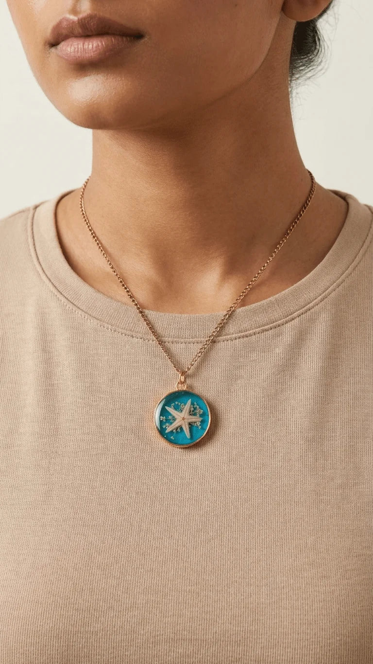A person wears a round gold necklace with a blue resin pendant featuring a white starfish and tiny shells.
