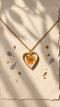 Heart-shaped resin pendant with a yellow flower and green leaf, on a gold chain, displayed on textured paper with petals.