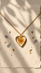 Heart-shaped resin pendant with a yellow flower and green leaf, on a gold chain, displayed on textured paper with petals.
