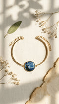 Gold-tone cuff bracelet featuring a circular resin charm with Van Gogh's Starry Night painting, on a light fabric background.