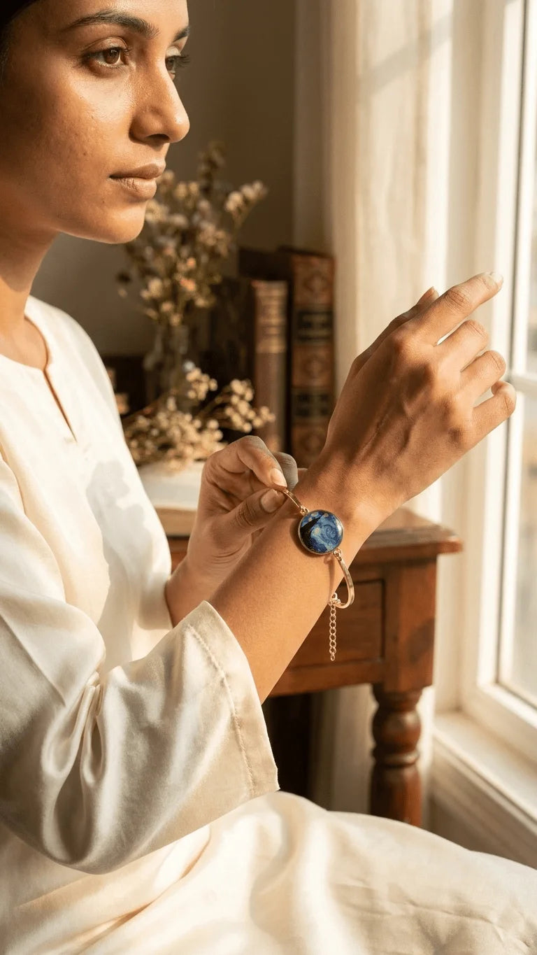 Woman in cream adjusting a gold bangle with a round, swirling deep blue resin charm on her wrist, by a sunlit window.