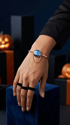 Hand wearing a gold bangle with a blue galaxy resin charm and black velvet rings, surrounded by Halloween decor.