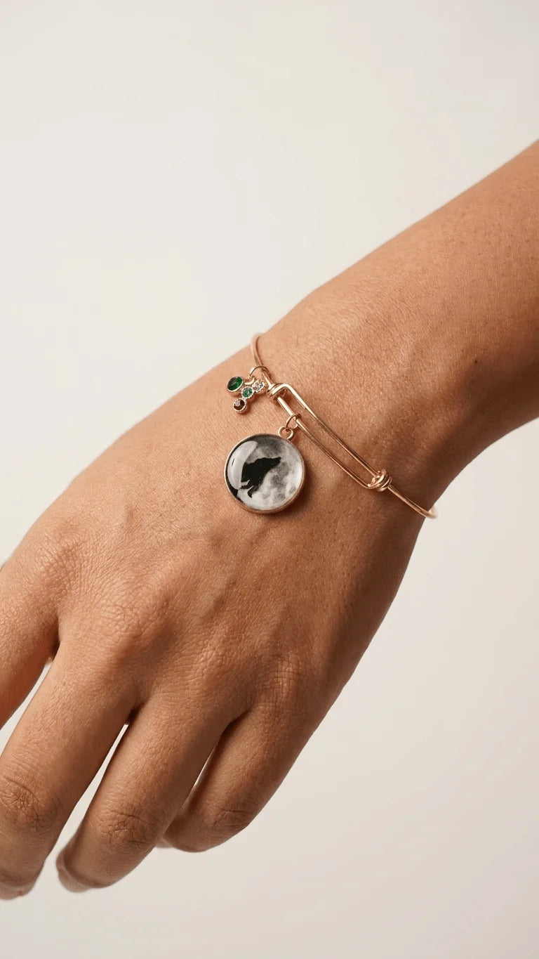 A person's hand wearing a rose gold bangle bracelet with a circular black cat and moon charm and a small green gemstone charm