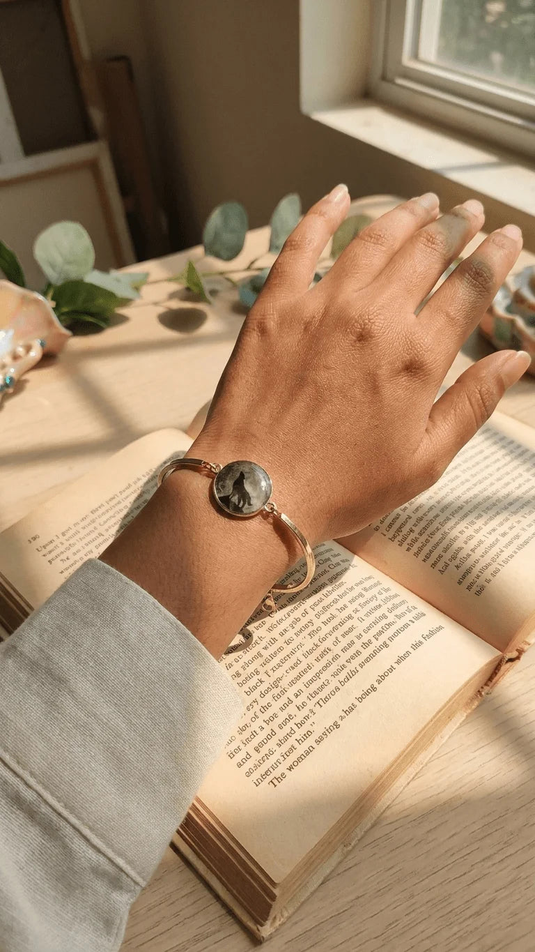 Hand wearing a gold bangle with a circular resin charm featuring a howling wolf silhouette, resting on an open book.