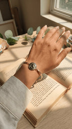 Hand wearing a gold bangle with a circular resin charm featuring a howling wolf silhouette, resting on an open book.