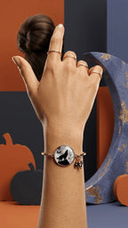 A hand wears a rose gold bangle featuring a howling wolf charm and black beads, against a dark blue and orange Halloween-them