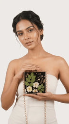 Woman holding a black resin clutch bag with embedded green leaves, white flowers, and gold flakes, with a gold chain strap.