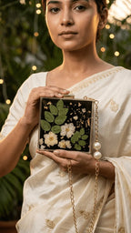 Woman in a cream saree holding a black resin clutch with white flowers, green leaves, gold flakes, and a pearl-accented gold