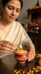 Woman in a cream saree lighting a yellow candle in a red decorated pot with a peacock feather, marigold petals on table.