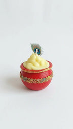 Red pot candle with yellow sculpted wax, gold trim, and a small peacock feather on a white background.