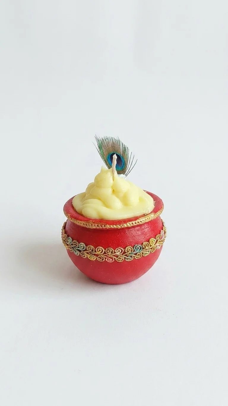 Red pot candle with yellow sculpted wax, gold trim, and a small peacock feather on a white background.