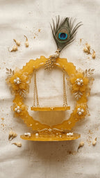 Golden resin jhula decor with embedded gold flakes, pearl-centered flowers, and a peacock feather, on white fabric with jasmi