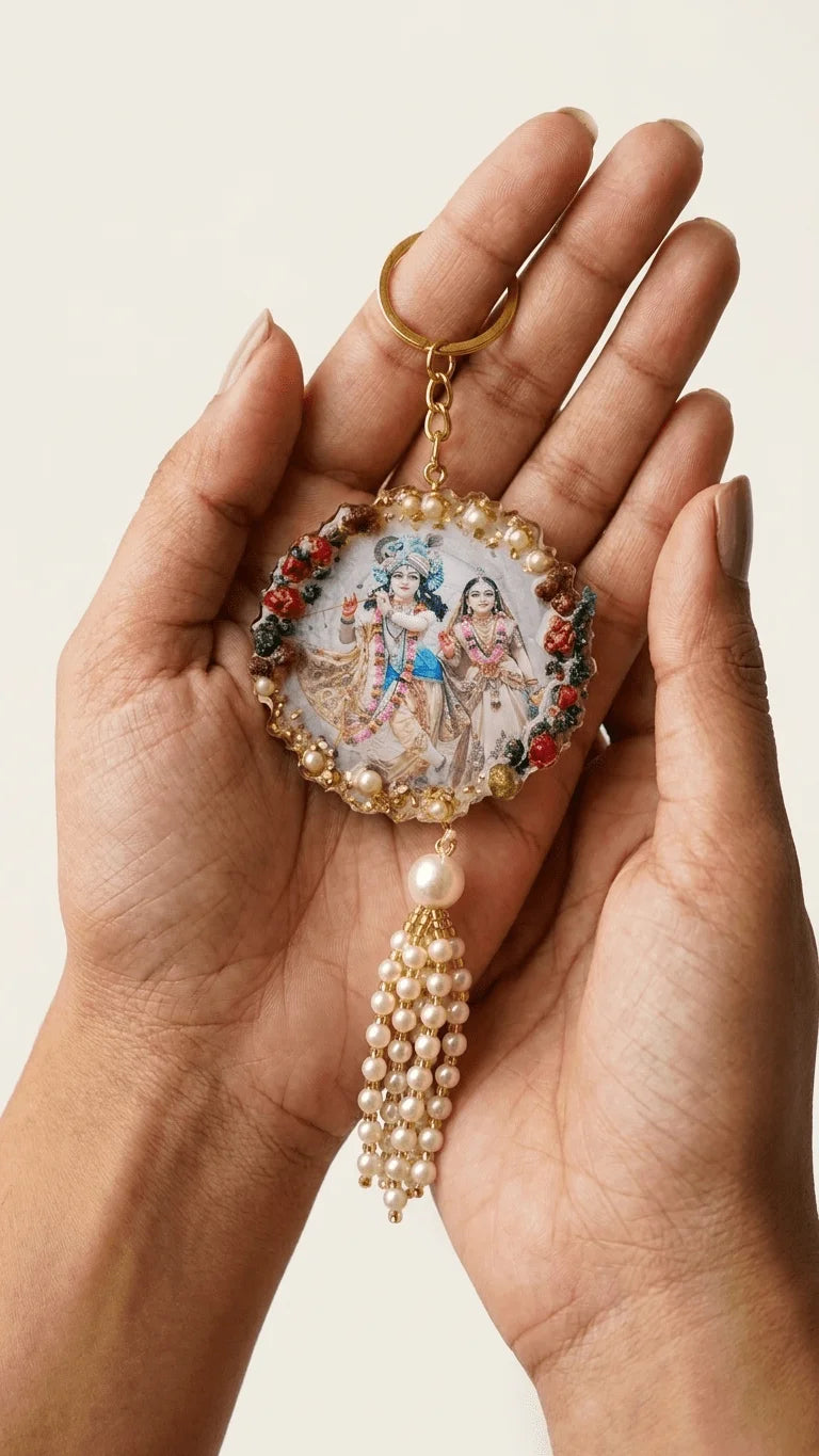 Hands holding a round resin keychain with Radha Krishna art, pearl border, and a pearl tassel.