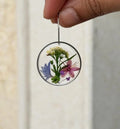 A hand holds a clear circular resin earring with pressed blue, pink, purple, and yellow flowers and green leaves inside.