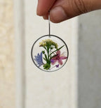 A hand holds a clear circular resin earring with pressed blue, pink, purple, and yellow flowers and green leaves inside.