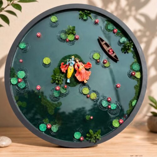 Circular resin art piece depicting Radha and Krishna standing on an island in a pond with lotus leaves, pink flowers, and a s