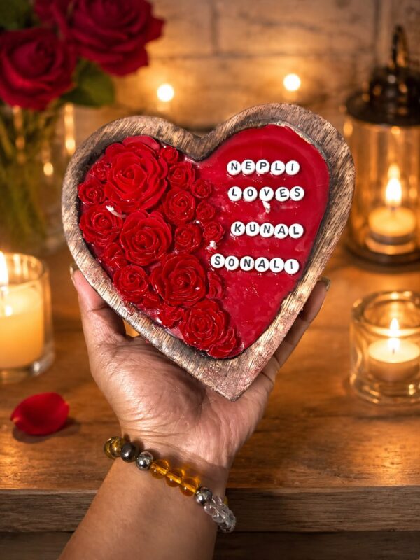 Hand holding a heart-shaped wooden tray with red resin roses and names 'NEPLI LOVES KUNAL SONALI' in a warm, candlelit settin