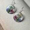 Two circular resin earrings with pressed blue, pink, yellow, and purple flowers inside, on an open book.