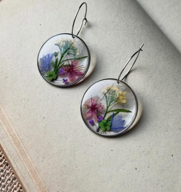Two circular resin earrings with pressed blue, pink, yellow, and purple flowers inside, on an open book.