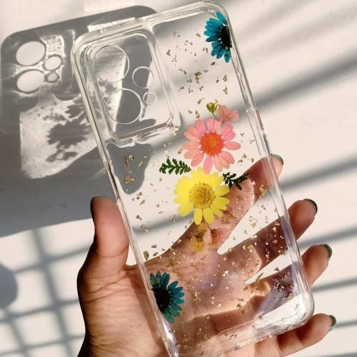 Hand holding a clear resin phone case with embedded blue, pink, and yellow dried flowers and gold flakes.