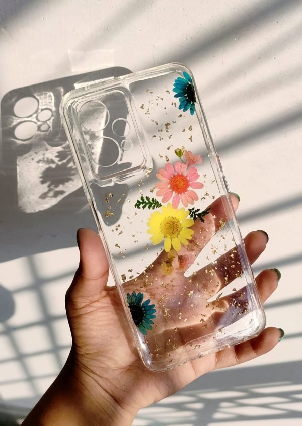 Hand holding a clear resin phone case with embedded blue, pink, and yellow dried flowers and gold flakes.