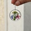 A hand holds a clear circular resin earring with pressed blue, pink, purple, and yellow flowers and green leaves inside.