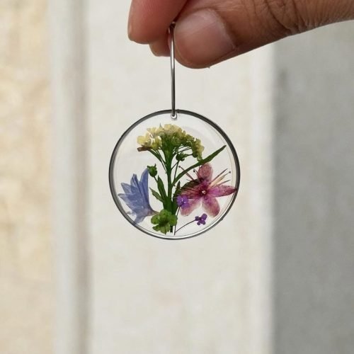 A hand holds a clear circular resin earring with pressed blue, pink, purple, and yellow flowers and green leaves inside.