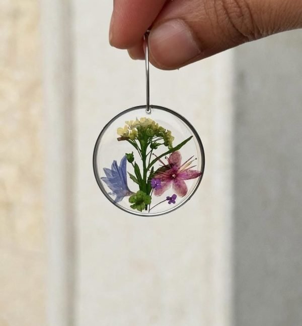 A hand holds a clear circular resin earring with pressed blue, pink, purple, and yellow flowers and green leaves inside.