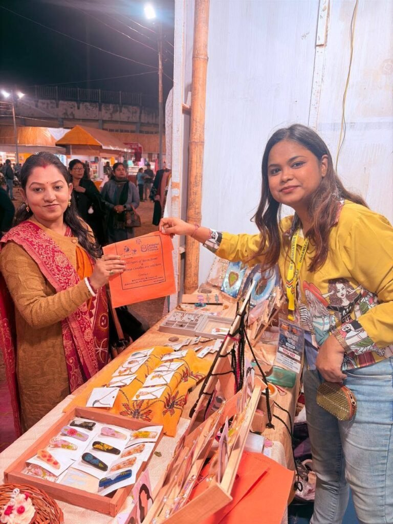 Two women at a market stall, one customer and one vendor, holding an orange flyer, with handmade resin jewelry and decor disp