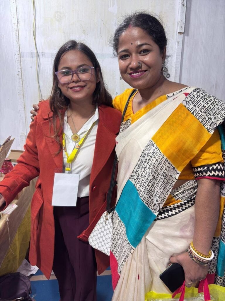 Two smiling women, one in an orange coat, the other in a white and yellow saree with patterned text, at an event.