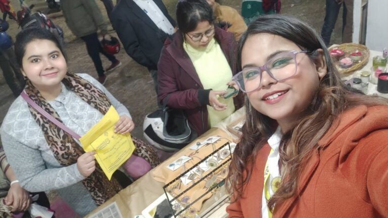 Three women at a market stall, with one smiling woman in an orange coat taking a selfie, and resin jewelry displayed.