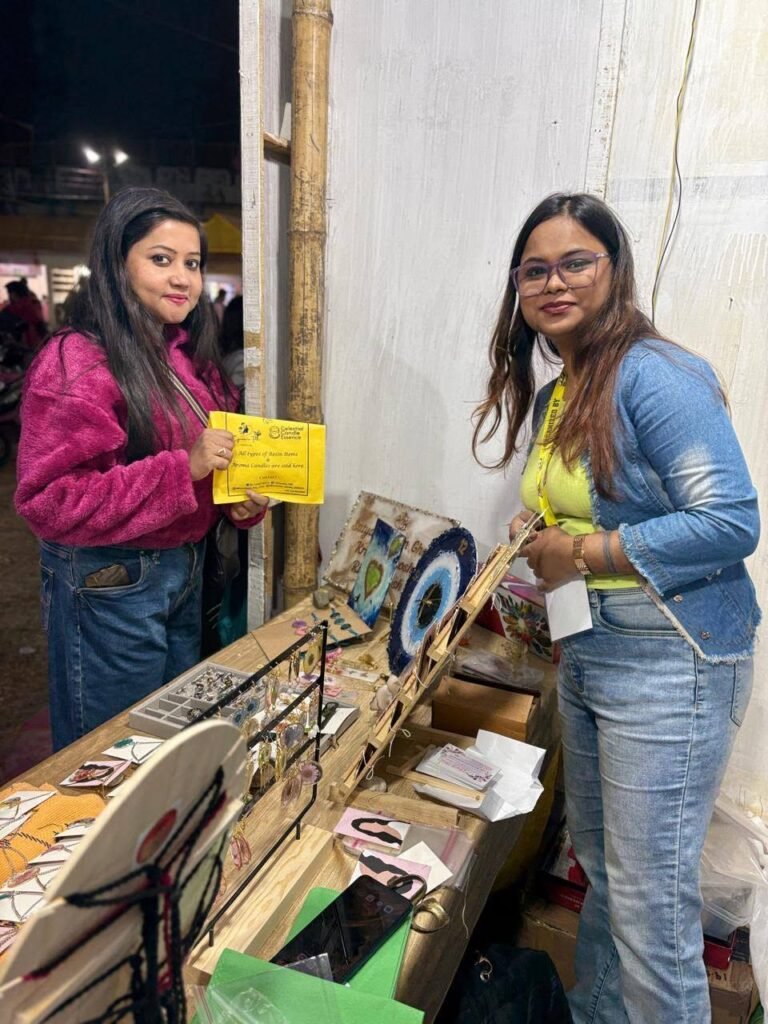 Two women at a market stall displaying handmade resin jewelry and decor, including earrings and an evil eye design.