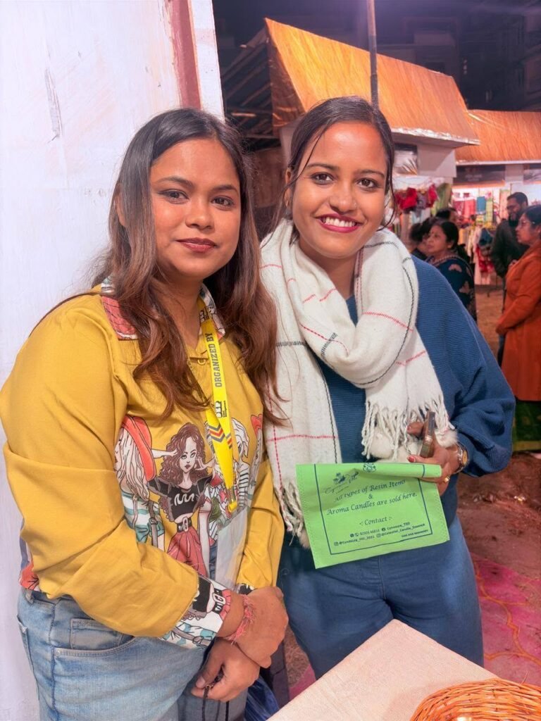 Two women smiling at an event, one wearing a yellow shirt and the other holding a green sign for resin items and candles.
