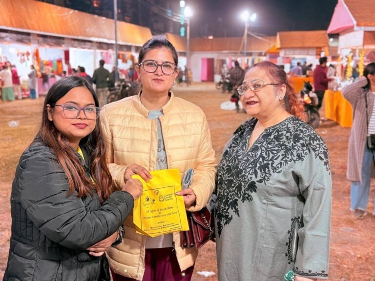 Three women smiling at an outdoor evening market, one holding a yellow bag from Cynosure Titli.