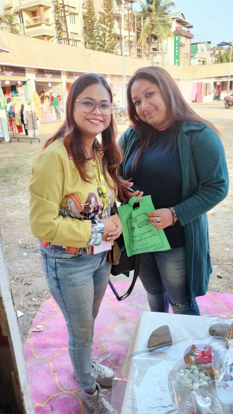 Two smiling women at an outdoor market stall. One wears a yellow jacket, the other a teal cardigan and holds a green bag.